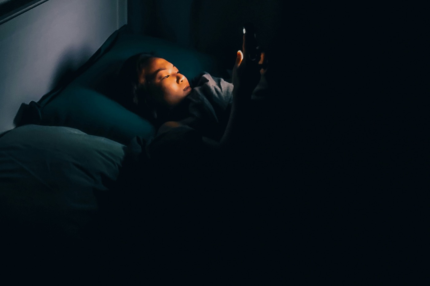 Persona acostada en la cama en un cuarto oscuro mirando el celular, con la pantalla como única luz