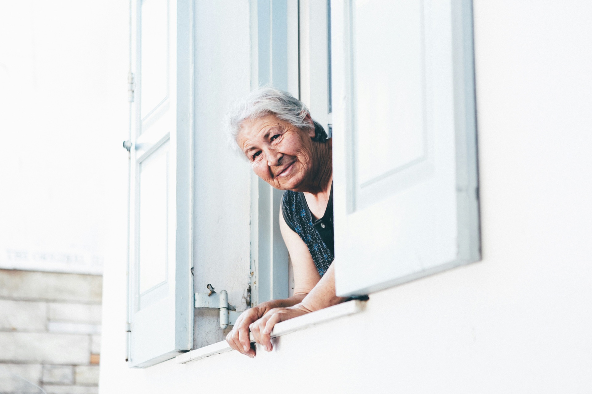 Una persona mayor sonriendo desde una ventana abierta con contraventanas blancas